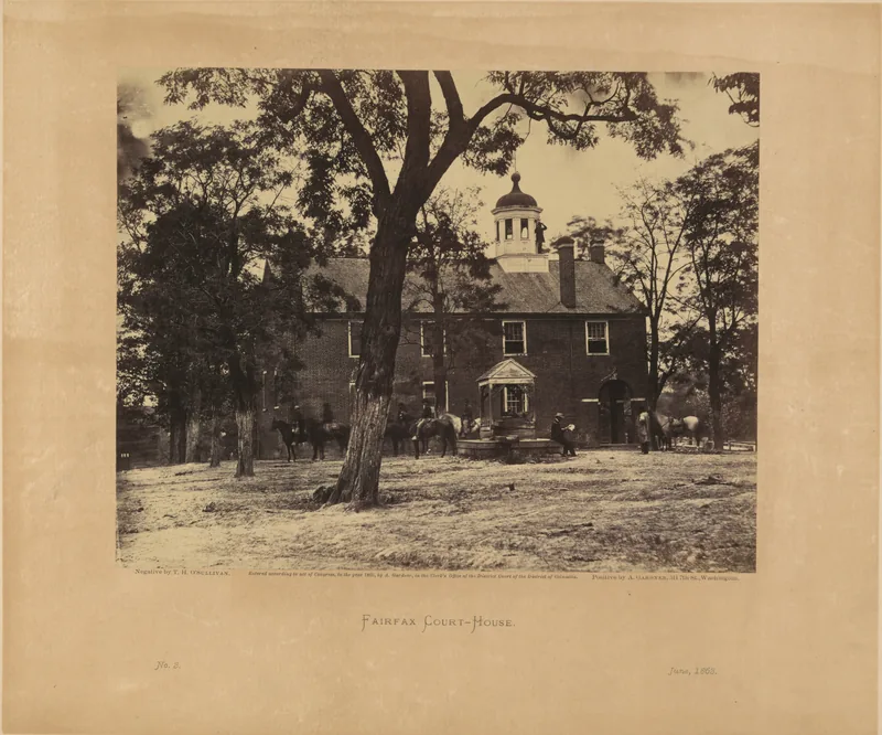 Sepia photograph of a two-storey brick courthouse with a cupola behind a large bare-trunked tree, with soldiers and saddled horses in the yard.